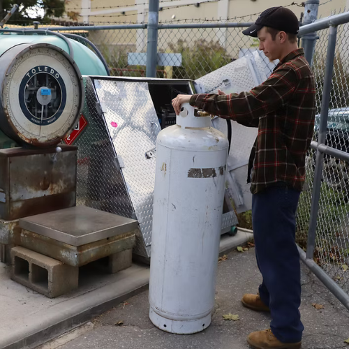 A person in a plaid shirt and cap stands outdoors, adjusting a large white gas cylinder. Nearby are industrial scales and metal panels, with a chain-link fence in the background. The scene conveys a work-focused atmosphere.