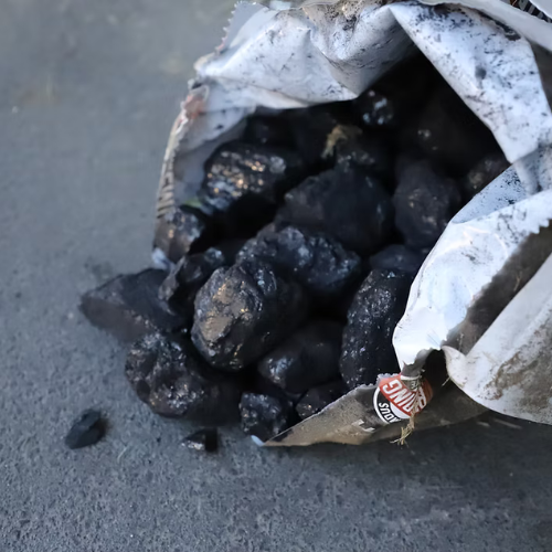 A torn paper bag spills shiny, black coal onto a gray surface. The coal pieces are chunky and irregular, contrasting with the smooth floor.