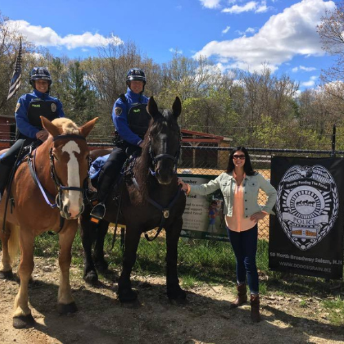 A woman stands next to two mounted police officers on horseback in front of a fenced area with trees and a blue sky in the background.