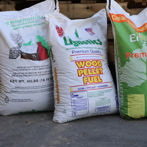 Three large bags of wood pellet fuel are standing upright on the ground. Each bag displays text and graphics related to the product. The setting appears industrial.