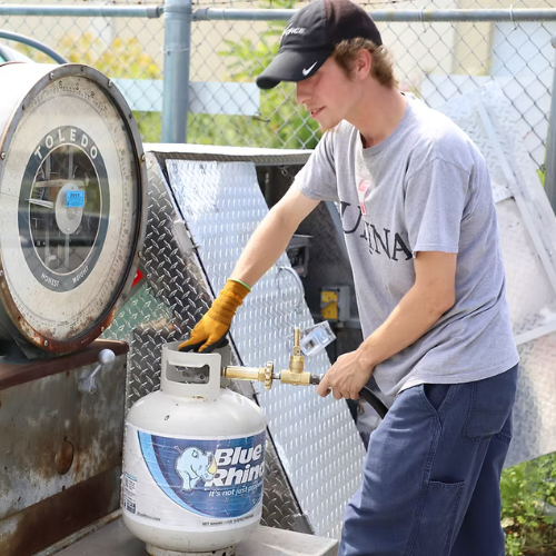 A person in a cap and gloves uses a scale to weigh a propane tank outdoors. They wear a gray shirt and blue pants, focused on their task.