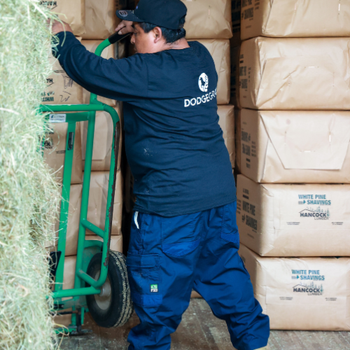 A worker maneuvers a green hand truck in a storage area, surrounded by large brown bags and hay bales. The scene conveys industry and diligence.