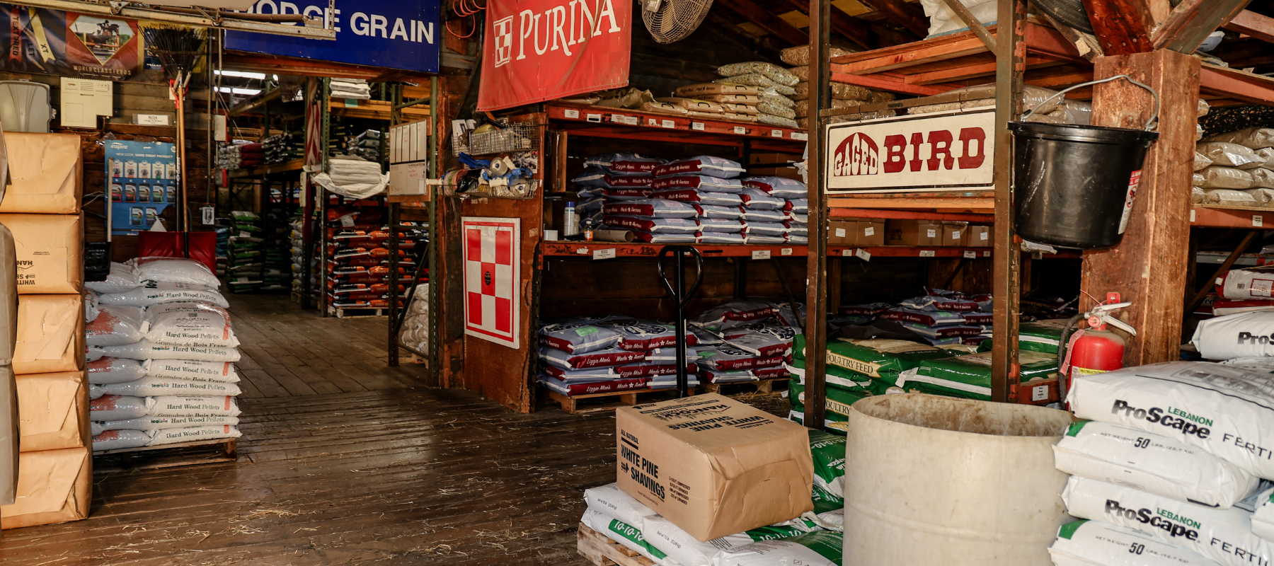 A view of an interior space resembling a store or warehouse, with shelves stocked with bags of grain, pet food, and fertilizers. The floor is wooden, and there are various signs hanging from the ceiling.
